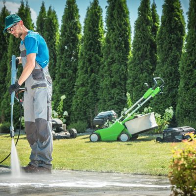 Caucasian Men in his 30s Cleaning Garden Paths and Driveway Using Professional Pressure Washer. Garden Equipment in the Background.